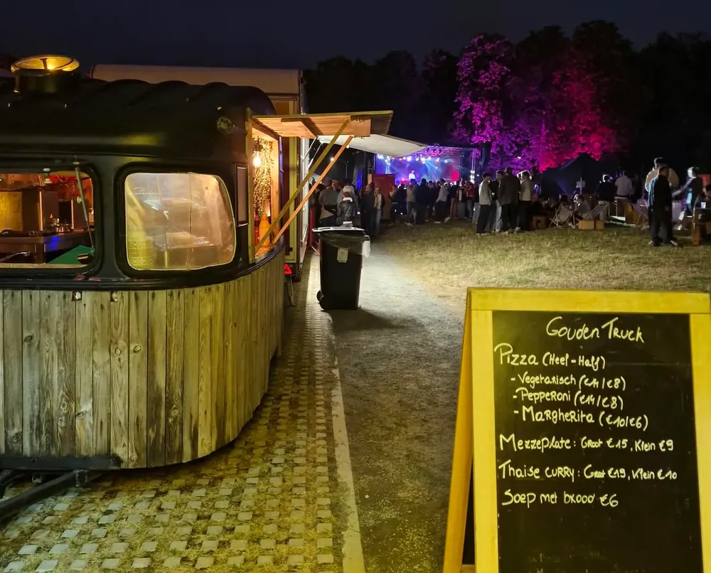 Een vintage houten caravan bij valavond naast een krijtbord, met feestgangers op de achtergrond